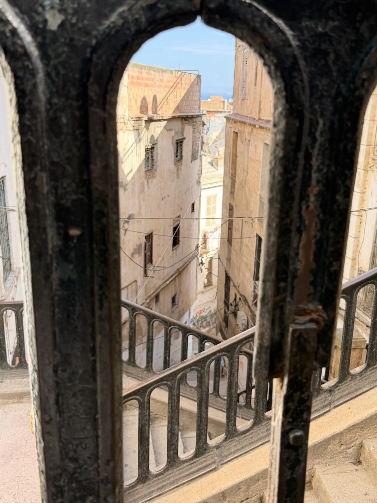 A stairway in the Casbah of Algiers seen through a weathered iron railing, with a glimpse of the sea beyond the old buildings